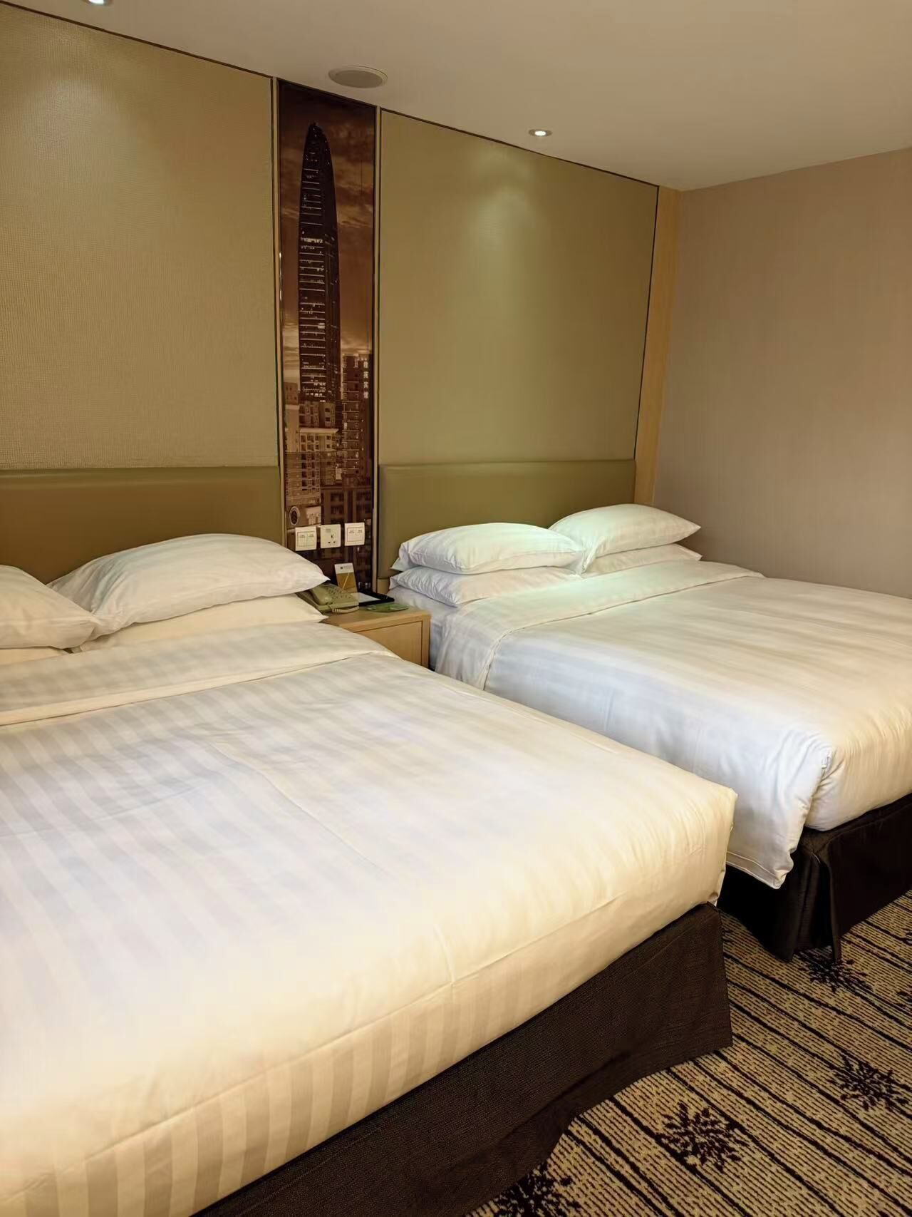 A hotel room with two beds, a large wall-mounted artwork of a cityscape, and a carpeted floor.