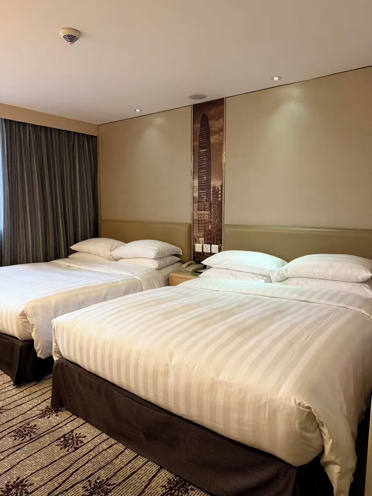 A hotel room with two beds, a large window with curtains, a wall-mounted artwork, and a carpeted floor.