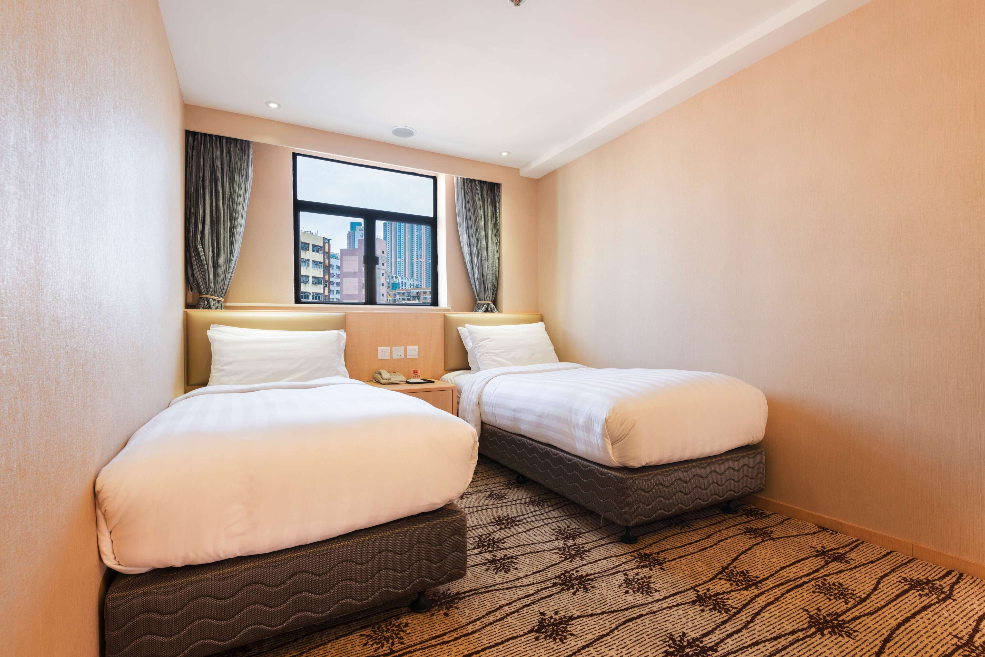 A hotel room with two beds, a large window with curtains, a patterned rug, and a nightstand with a lamp.