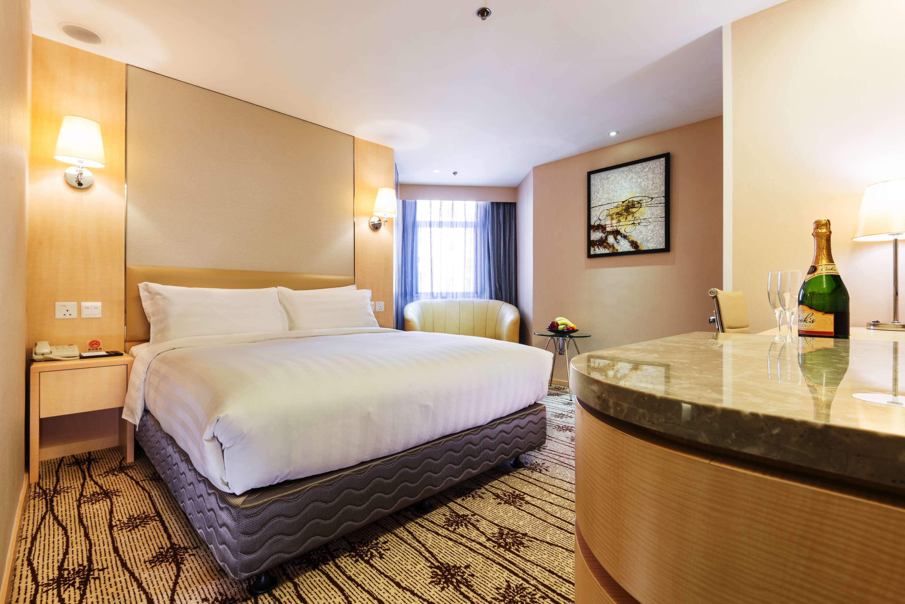Habitación de hotel con una cama grande, una mesita de noche con lámpara, una pequeña mesa redonda con una botella de champán y una silla.