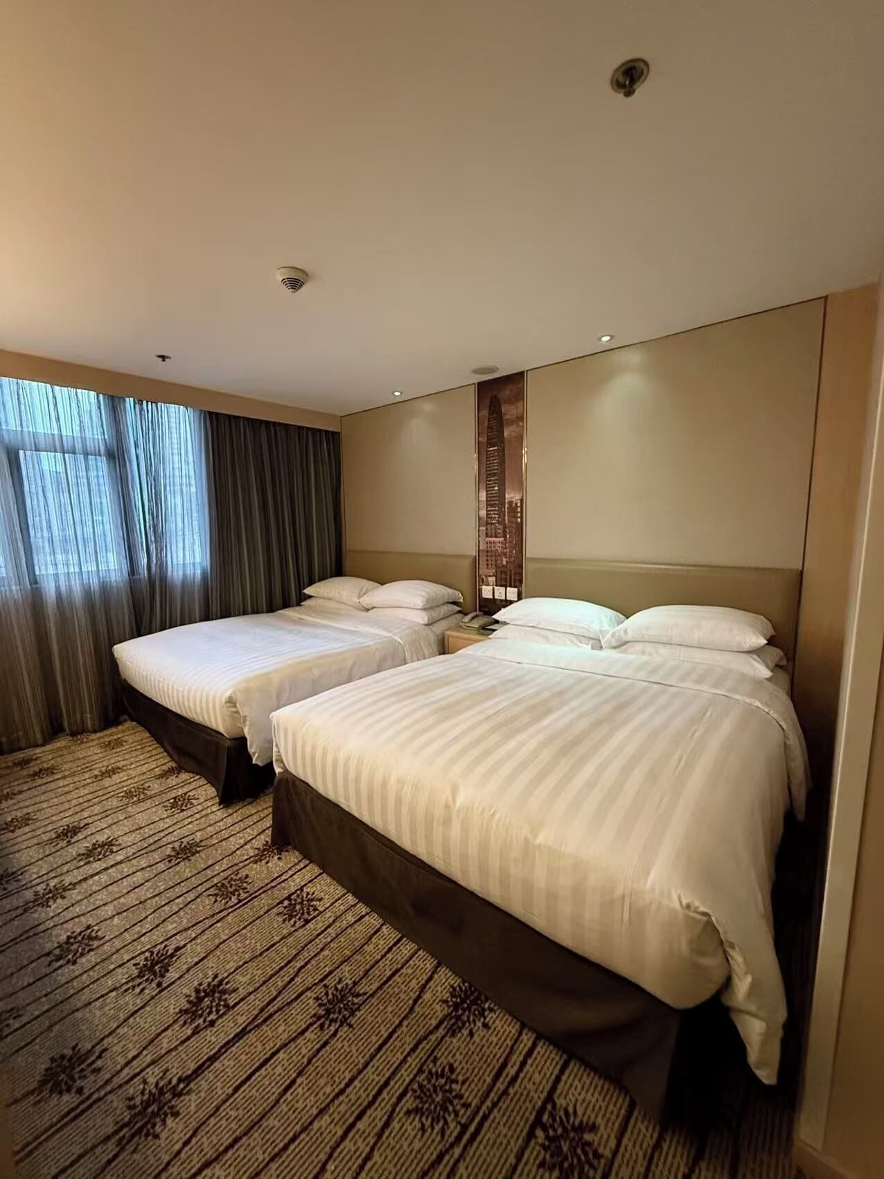 Ein Hotelzimmer mit zwei Betten, einem großen Fenster mit Vorhängen, einem gemusterten Teppich und einem an der Wand befestigten Kunstwerk.