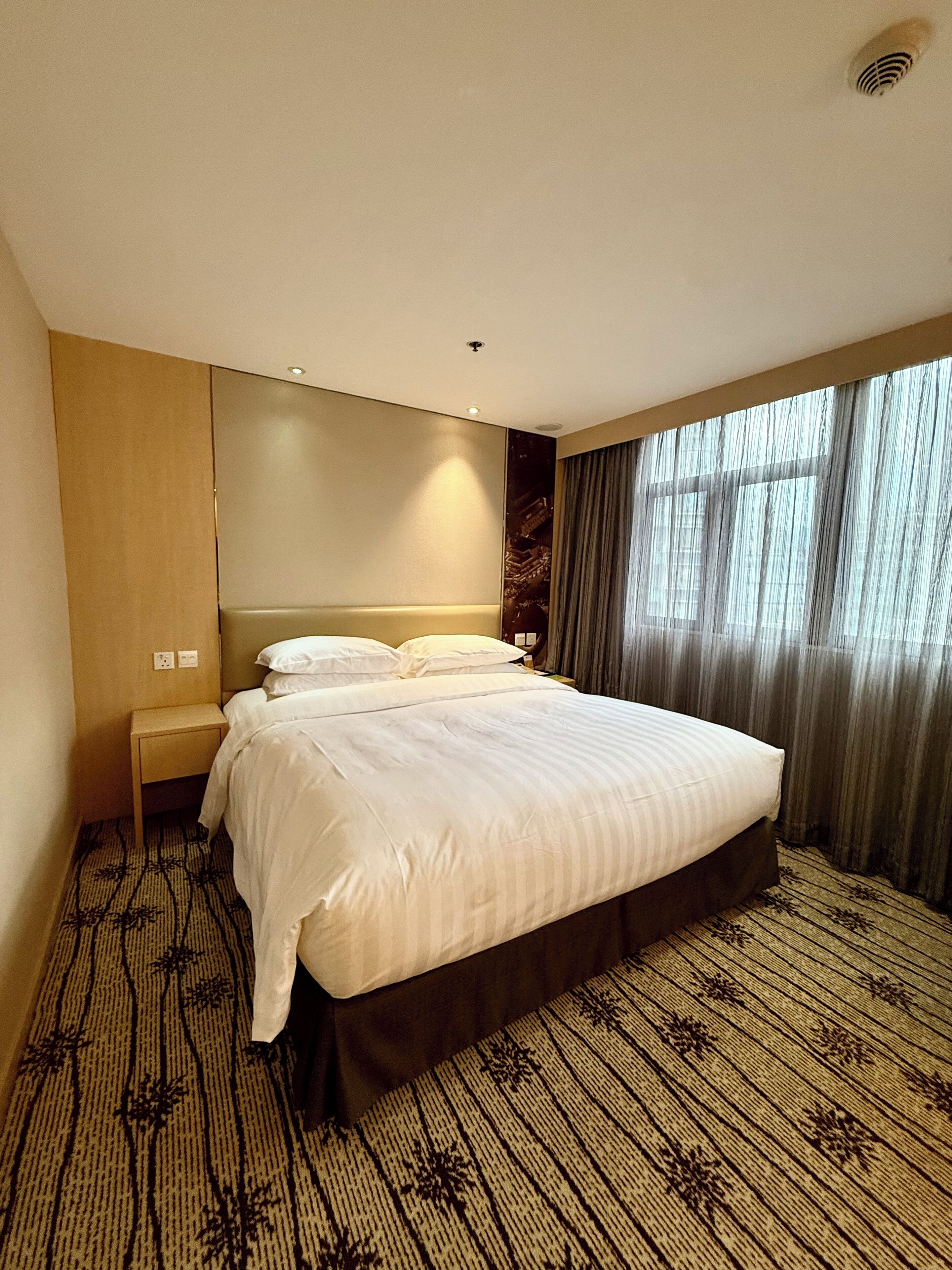 Habitación de hotel con una cama grande, una mesita de noche, un televisor montado en la pared y una ventana con cortinas.