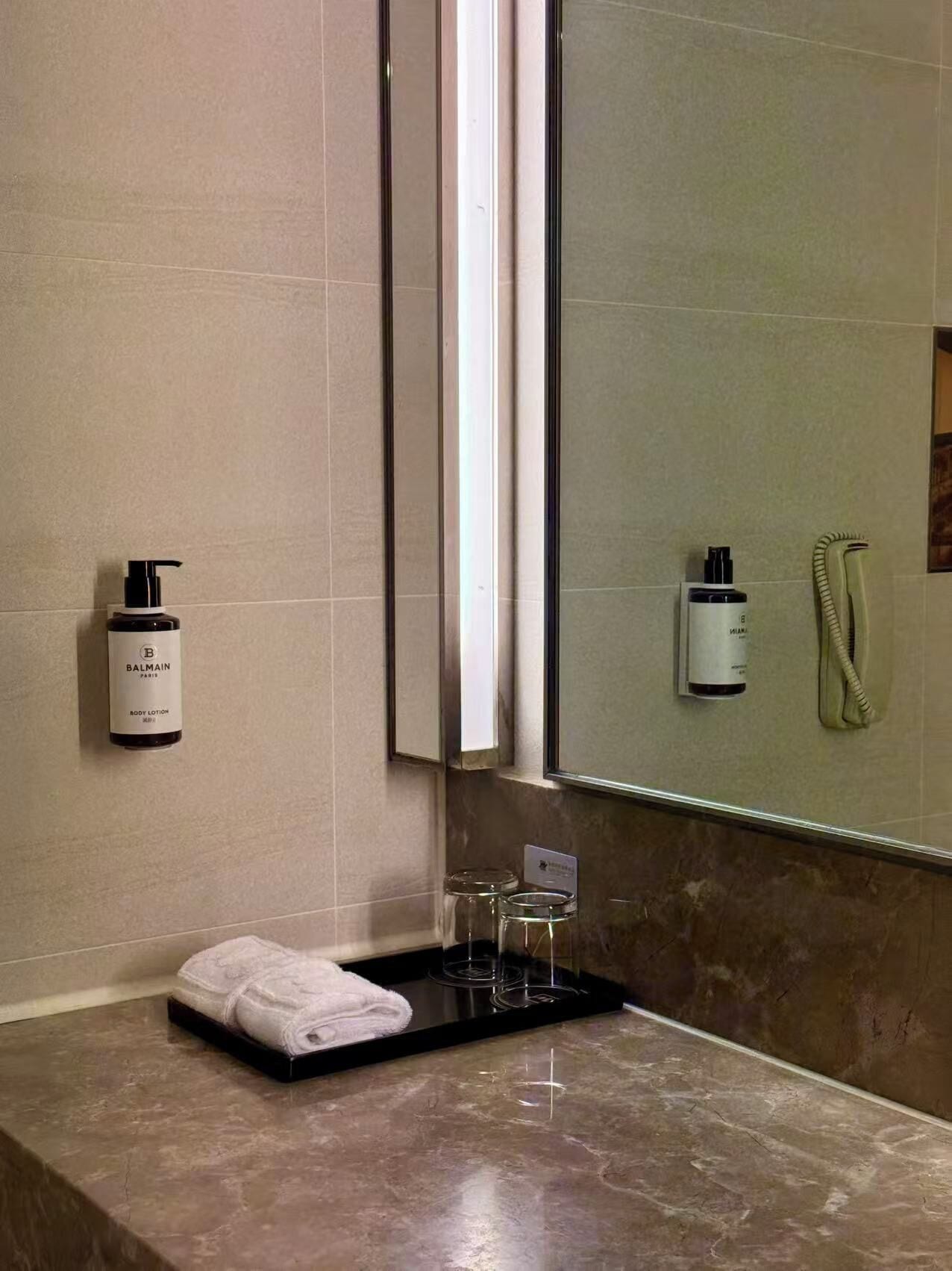 Un baño con encimera de mármol, una toalla blanca, una botella de loción corporal Balmain, un secador de pelo y un teléfono.