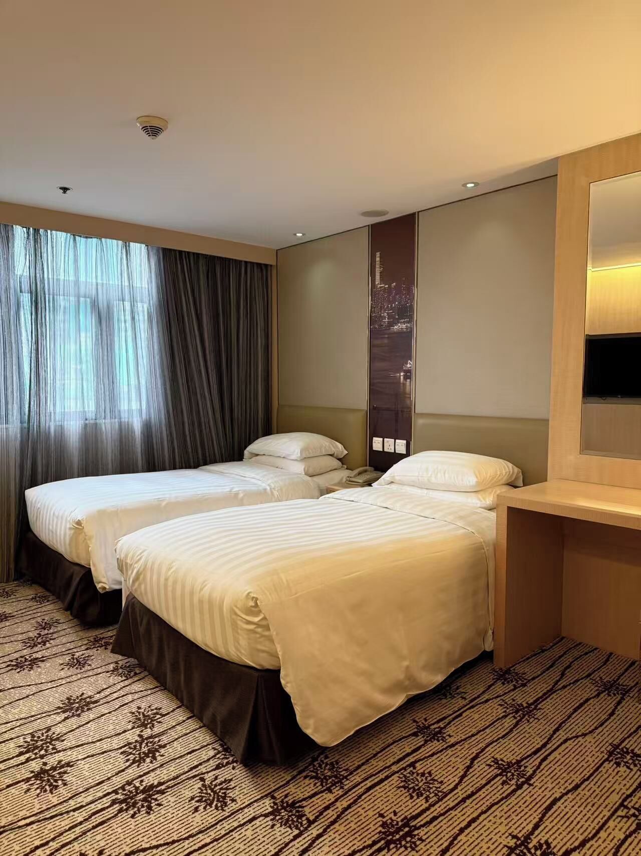 Une chambre d’hôtel avec deux lits, une grande fenêtre avec des rideaux, une vue sur la ville et une table de chevet en bois.