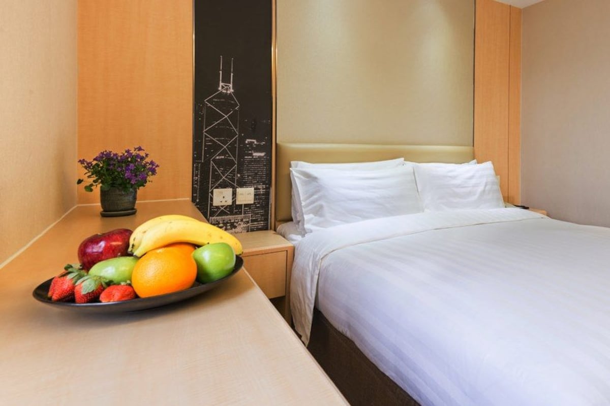 A hotel room with a bed, a nightstand with a fruit bowl, a wall-mounted clock, and a door.