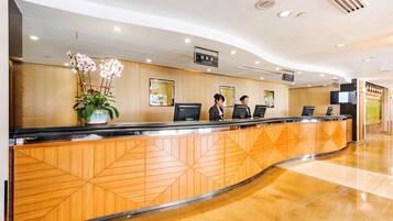 A hotel reception area with a curved wooden reception desk, two receptionists, a potted orchid, and a sign indicating the cashier area.