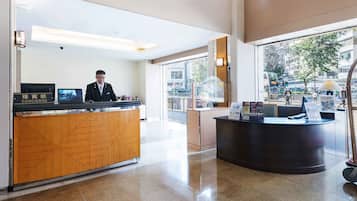 A hotel reception area with a receptionist, a wooden reception desk, a black counter, and a view of the street outside.