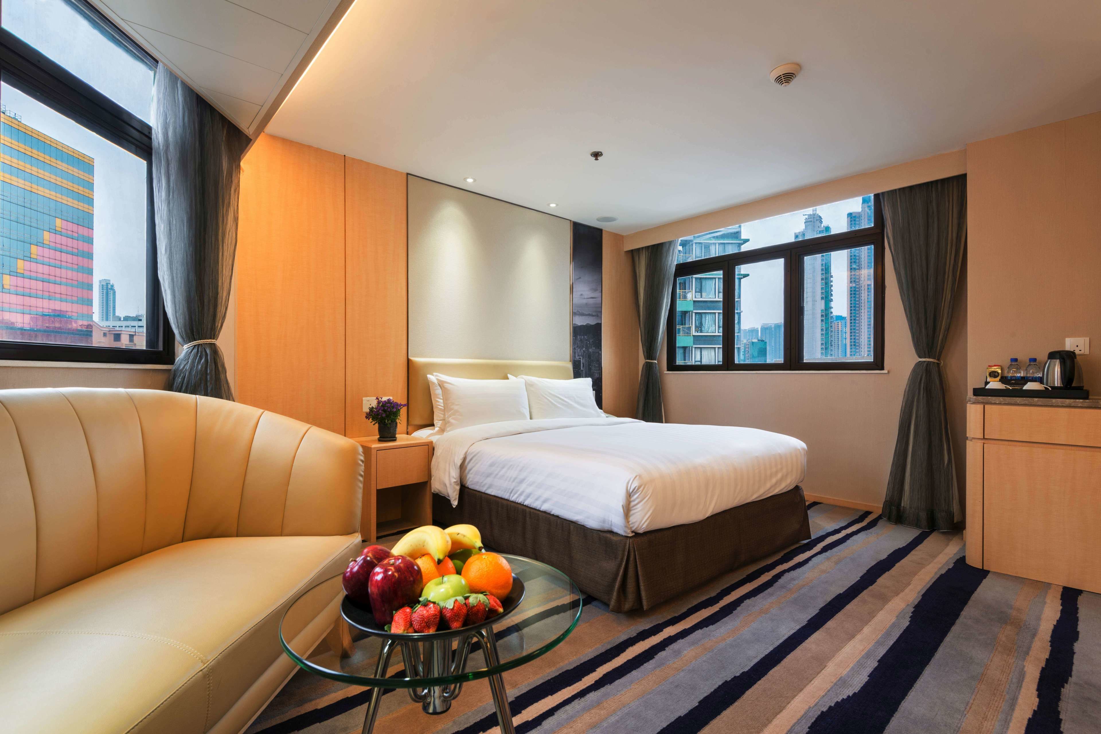 A hotel room with a bed, a sofa, a glass table with fruit, a view of the city, and a desk with a coffee maker.