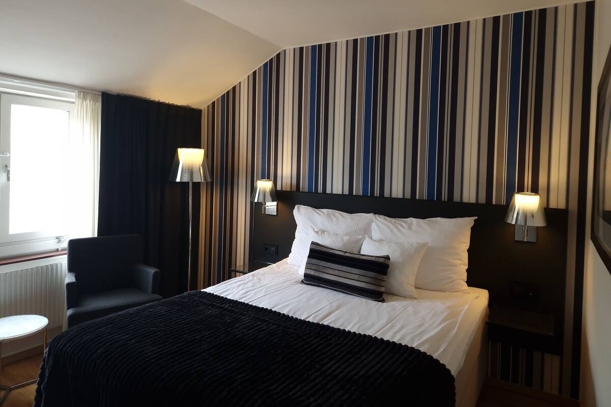Quarto de hotel com parede listrada, cama com almofadas brancas e listradas, uma cadeira preta e duas luminárias de cabeceira.