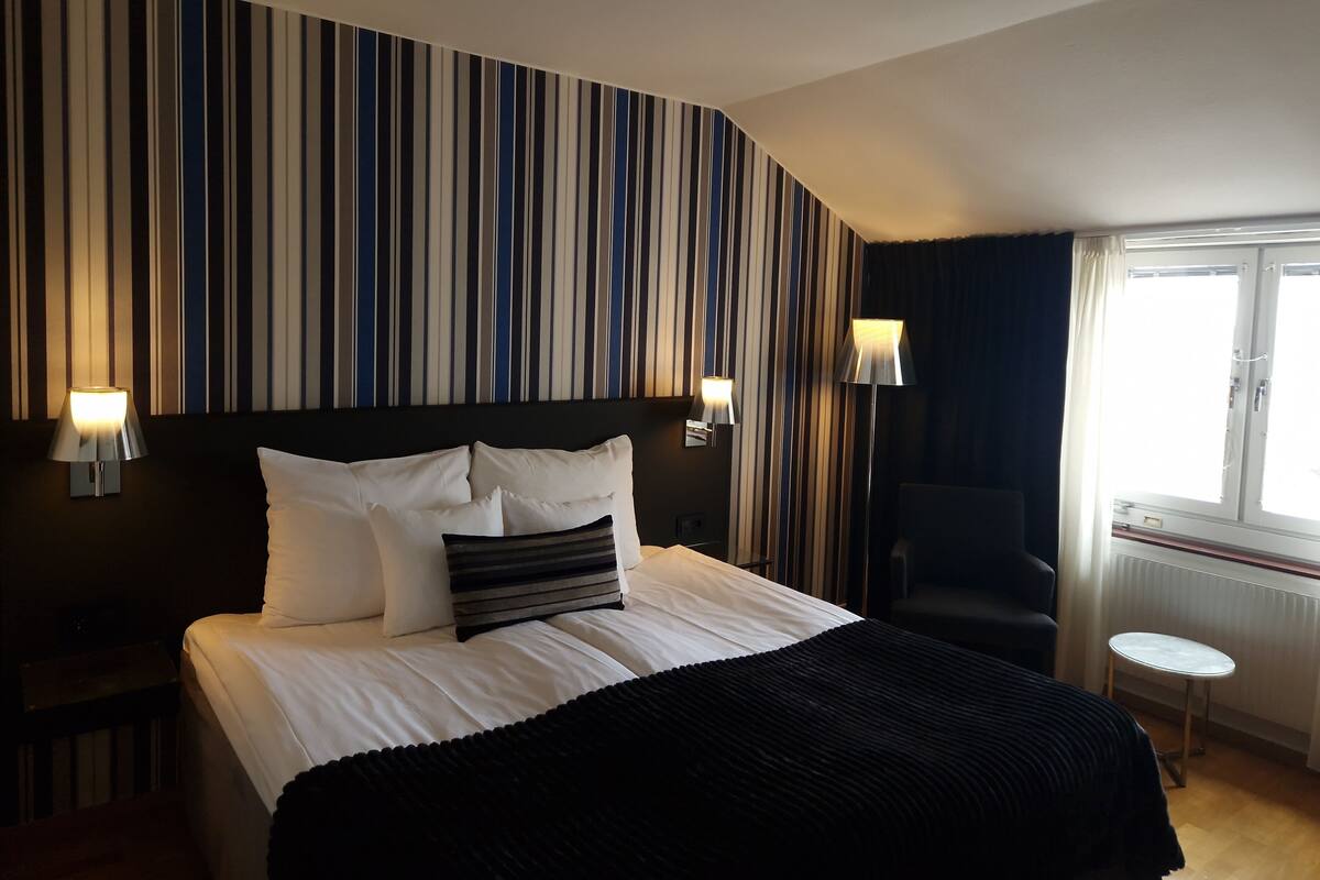 Quarto de hotel com parede listrada, cama com roupa de cama preta e branca, duas luminárias de cabeceira, uma cadeira e uma pequena mesa junto à janela.