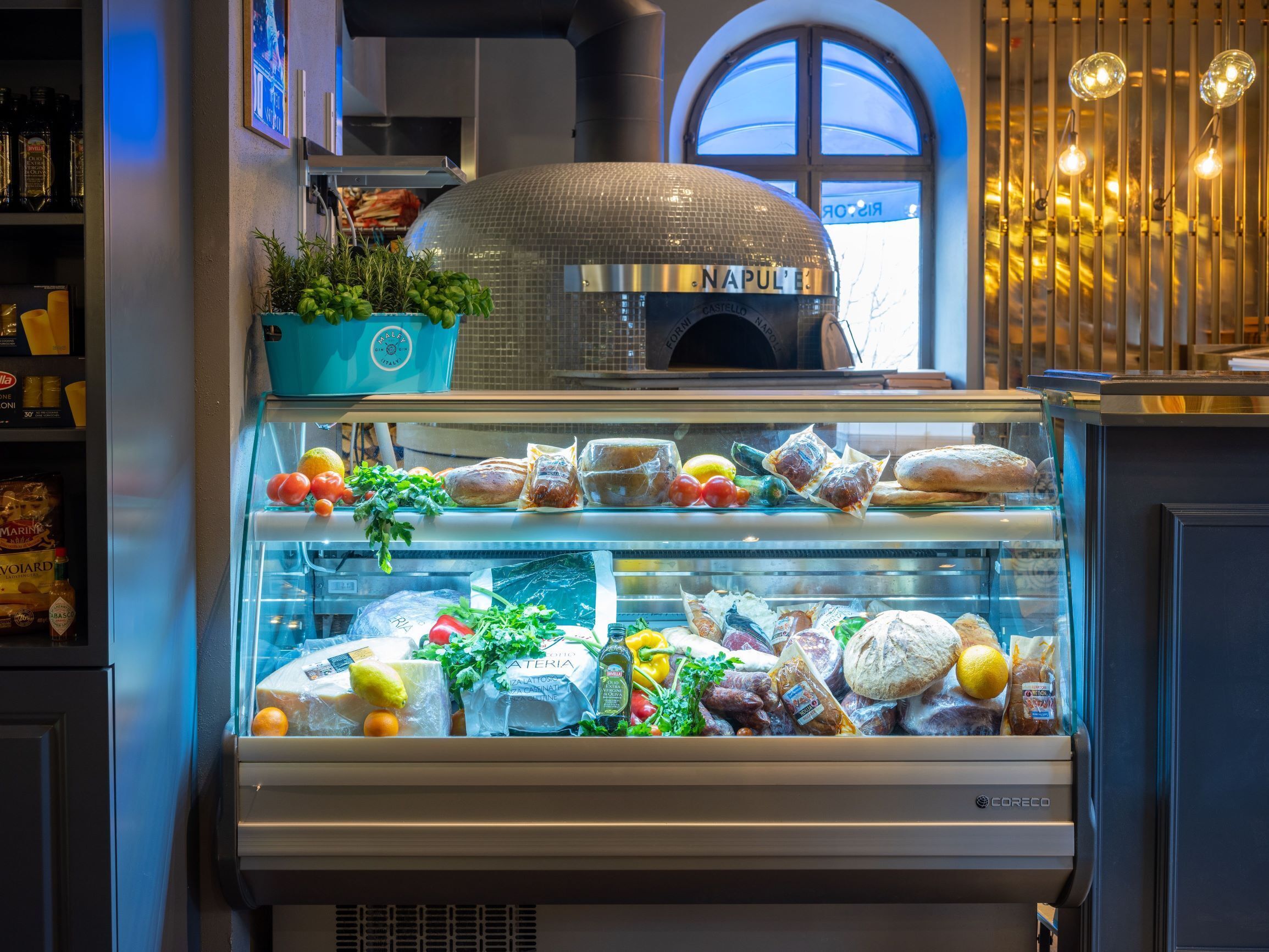 A bakery display case with various breads and pastries, a potted plant, and a pizza oven in the background.