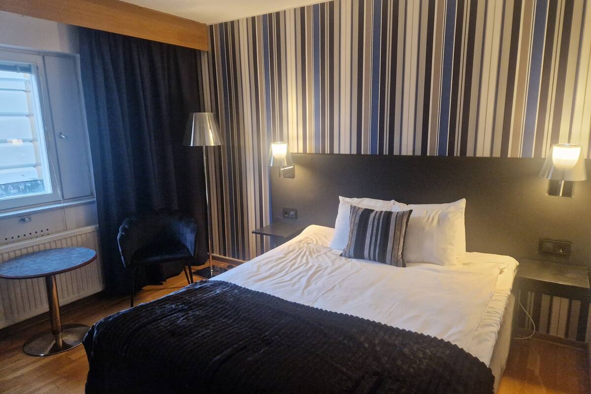 Quarto de hotel com cama, poltrona, mesinha e janela.