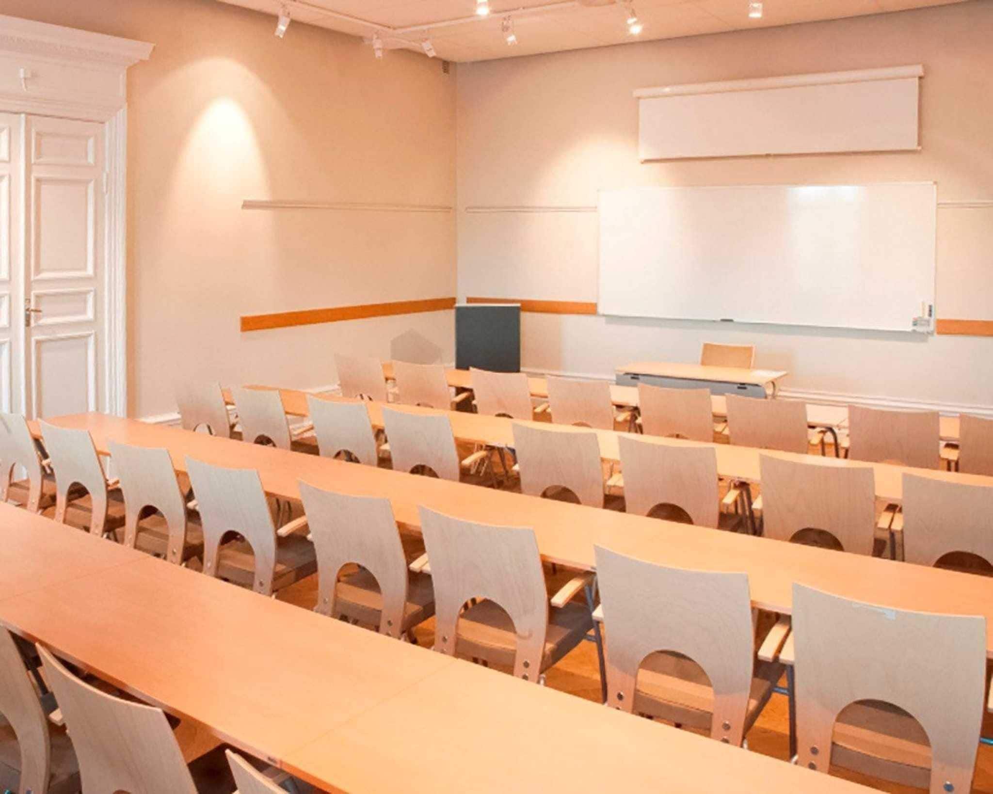 Una sala de conferencias con mesas largas y sillas, una pizarra y una pantalla para proyector.