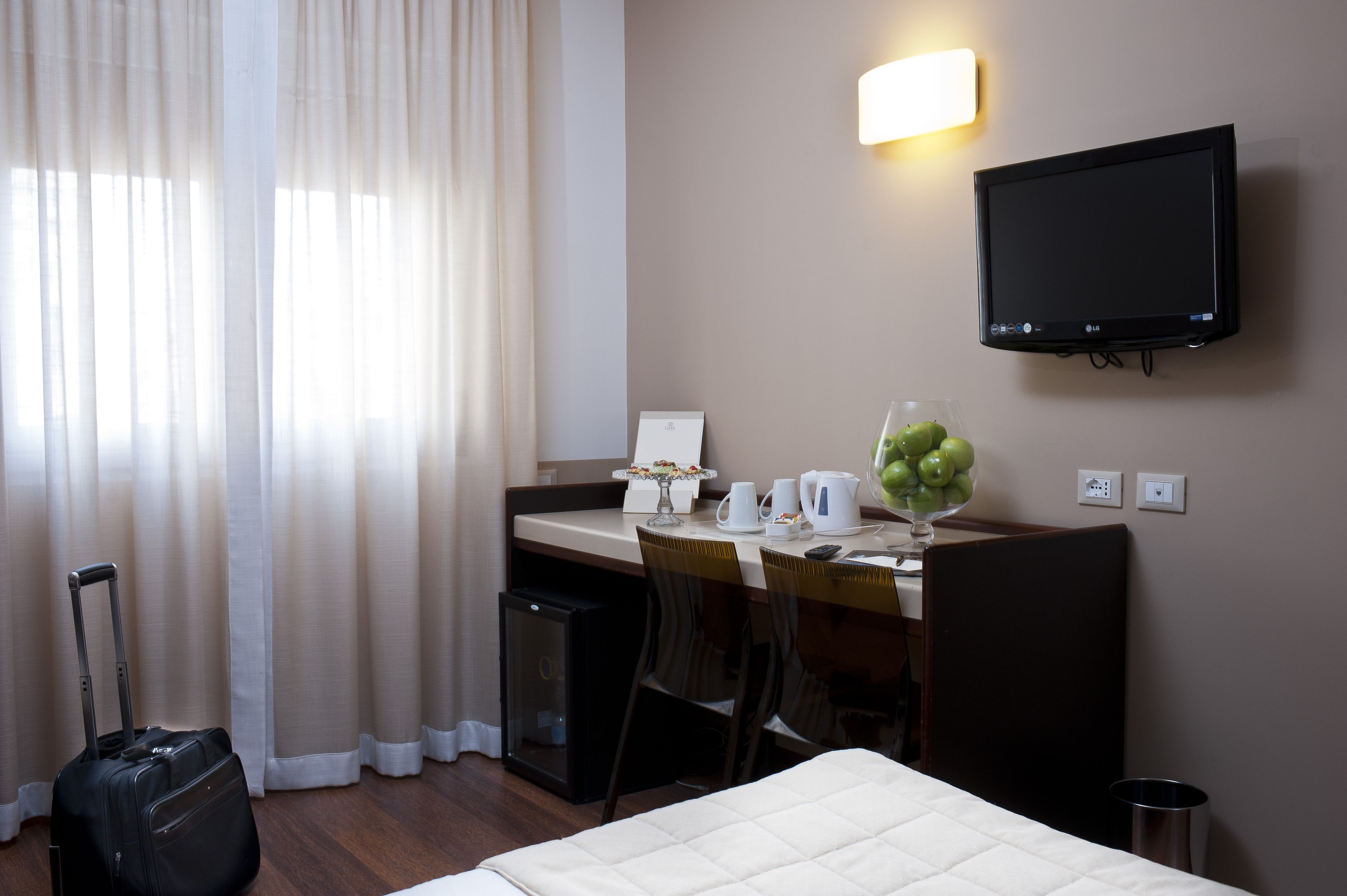 A hotel room with a bed, a desk with a computer, a TV, a small fridge, and a suitcase.