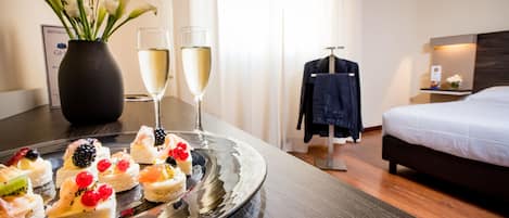A hotel room with a dining table set with a variety of desserts, two glasses of champagne, and a vase of flowers.