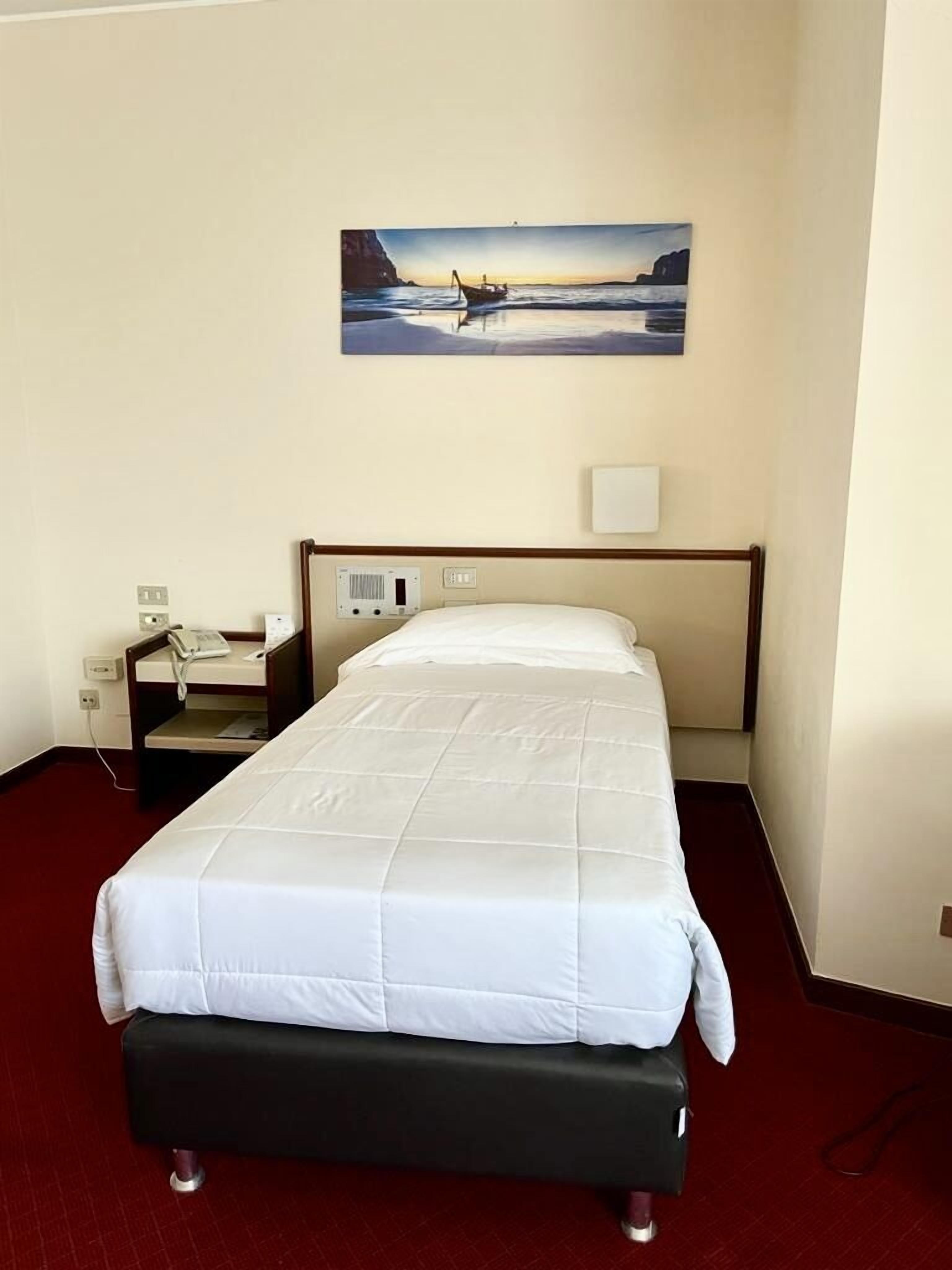 Une chambre d’hôtel avec un lit, une table de chevet, un téléphone, un tableau représentant un bateau sur l’eau et une moquette rouge.
