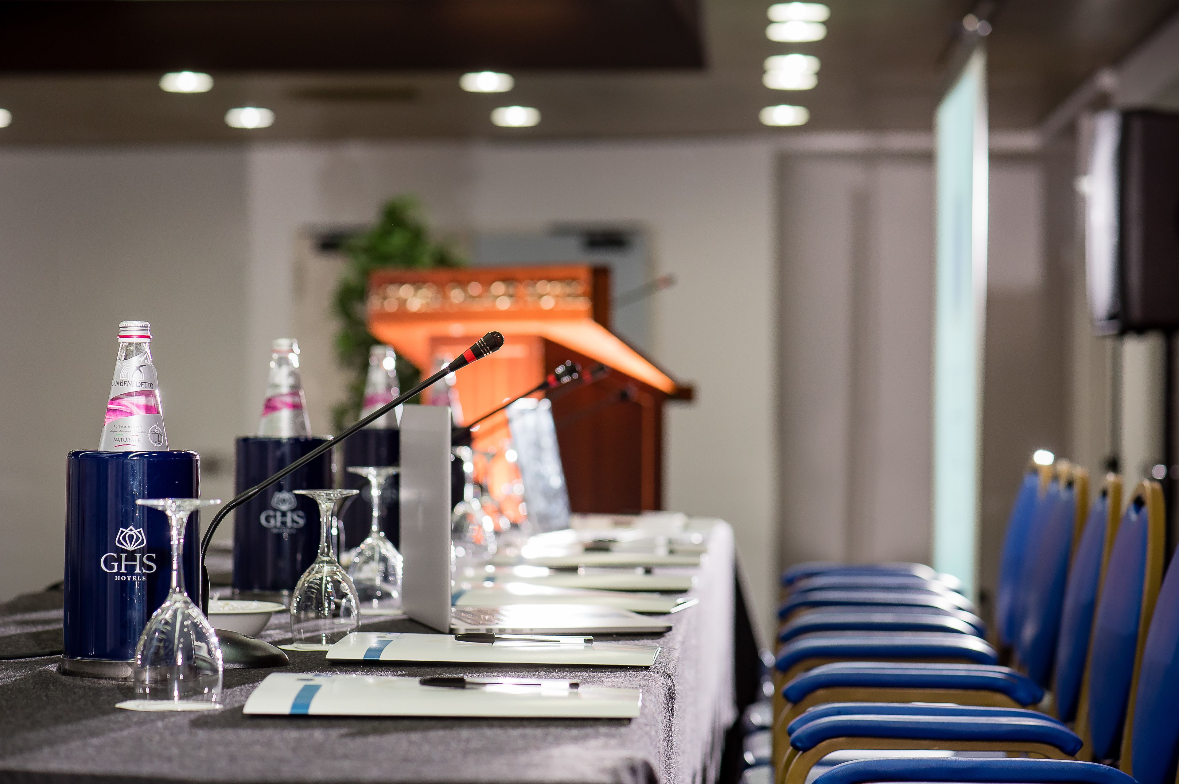 A conference table set with GHS branded bottles, wine glasses, and microphones, with blue chairs along the side.