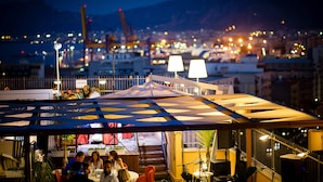 Un espace extérieur aménagé pour les repas, avec des tables et des chaises, un coin salon abrité et une vue sur le paysage urbain, avec des bâtiments et une grue en arrière-plan.