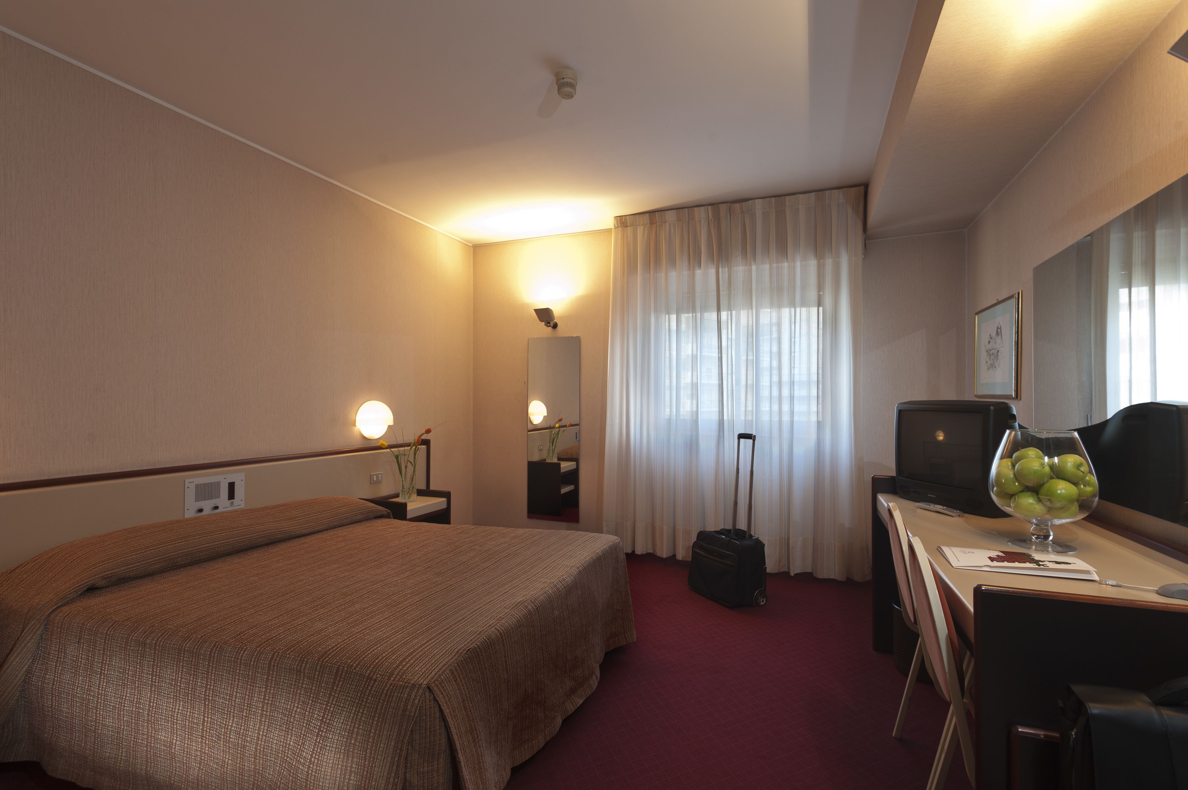 A hotel room with a bed, a desk with a computer, a suitcase, and a bowl of fruit.