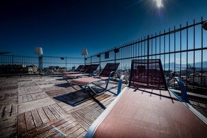 A rooftop terrace with sun loungers and a railing, offering a view of the cityscape and mountains.