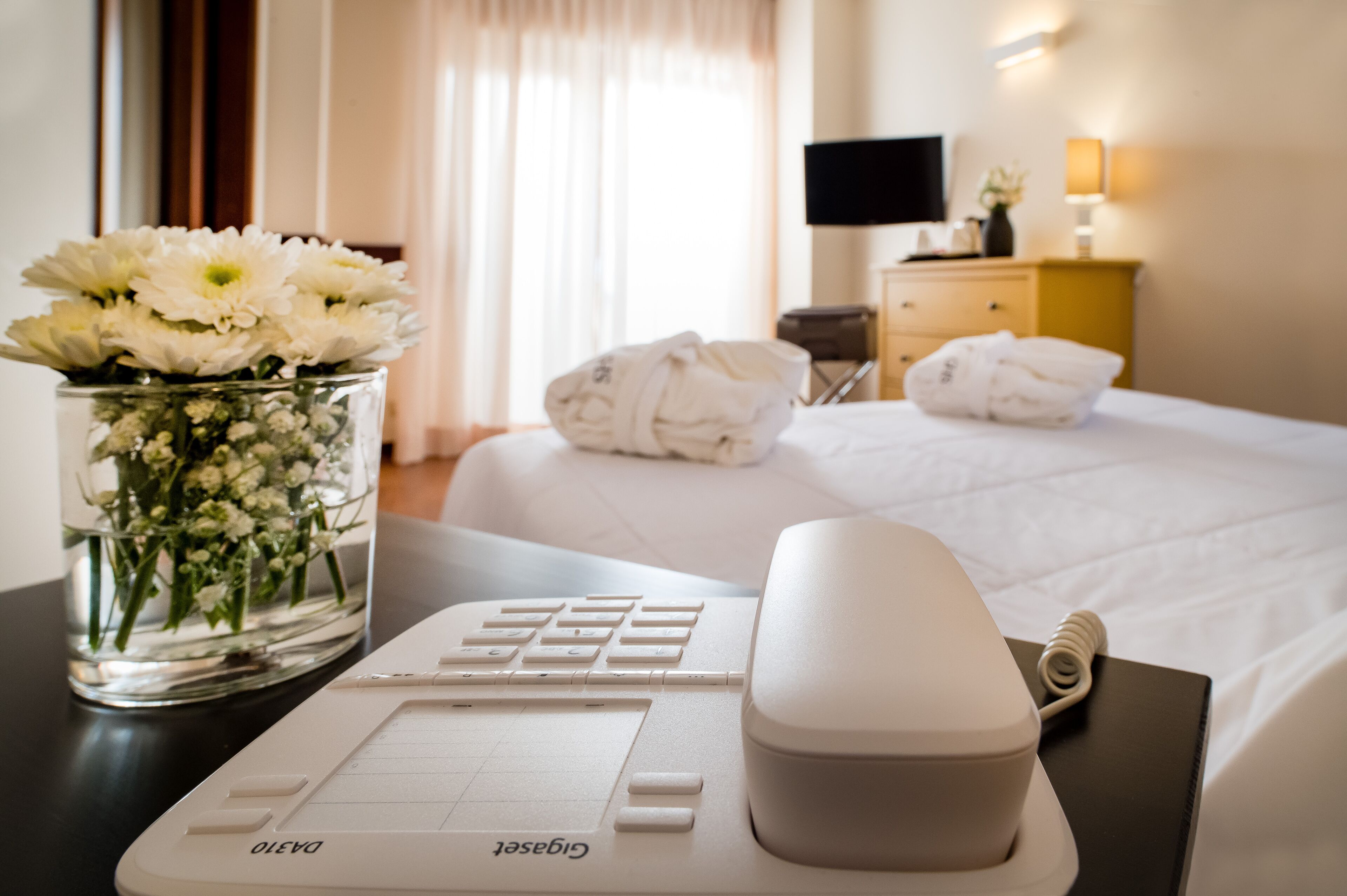 Una habitación de hotel con una cama, un escritorio con computadora, un jarrón de flores y un televisor en la pared.