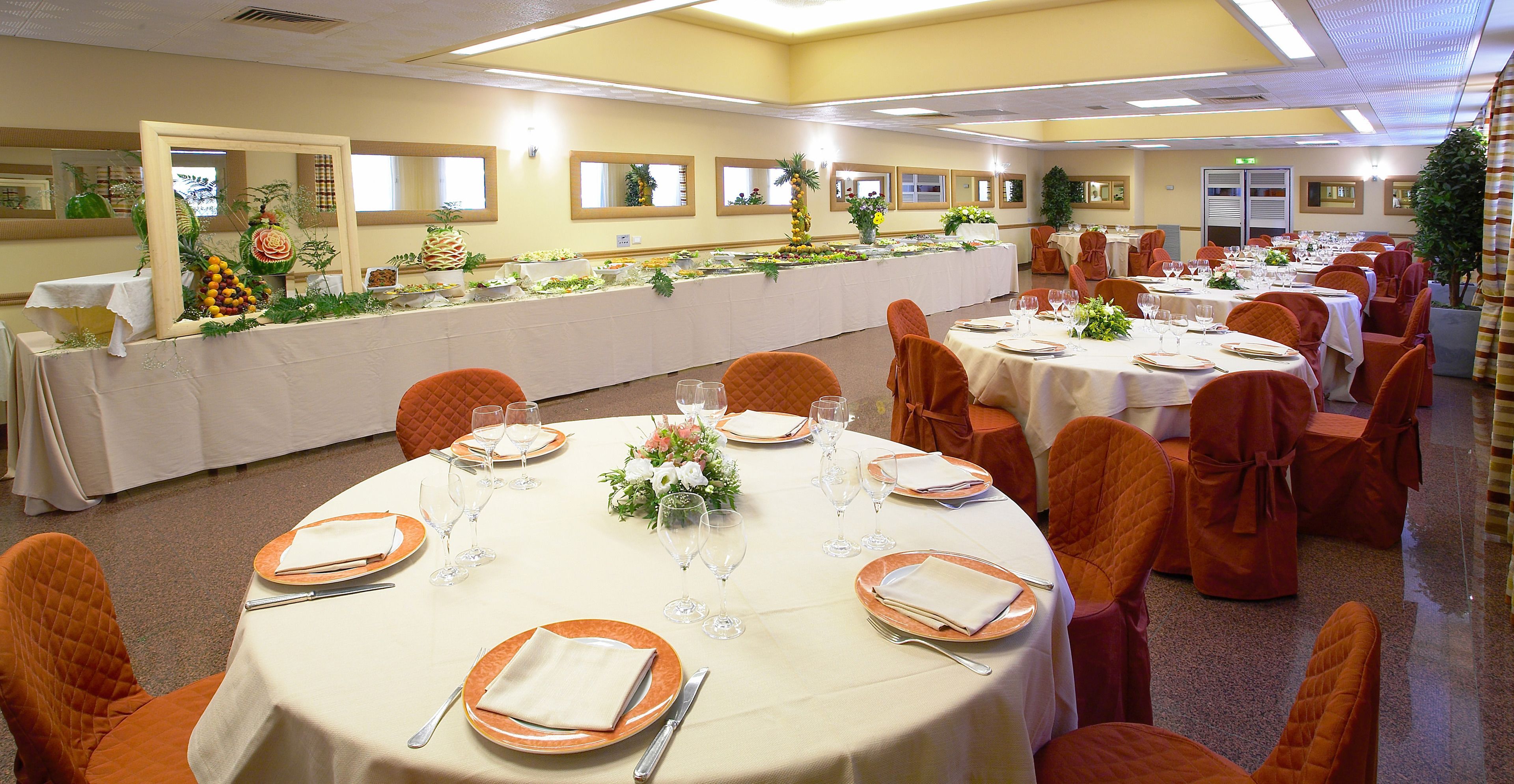 A banquet hall with round tables set for dining, orange chairs, and a long buffet table with food and decorations.