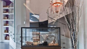 Un intérieur moderne avec une vitrine en verre contenant des livres et des objets décoratifs, un escalier au design original et un grand vase orné de branches.