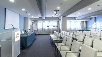 Ein Konferenzraum mit Reihen weißer Stühle, einem Rednerpult und einer Präsentationsleinwand.