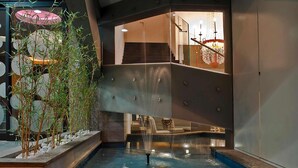 Un espace piscine intérieur moderne, doté d’un élément décoratif aquatique, de murs en verre et d’un escalier menant à un niveau supérieur.