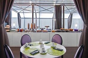A dining area with a high ceiling and large windows, featuring a buffet setup with various food items and a table set for four with green placemats. - Hotel Caravel (Rome)