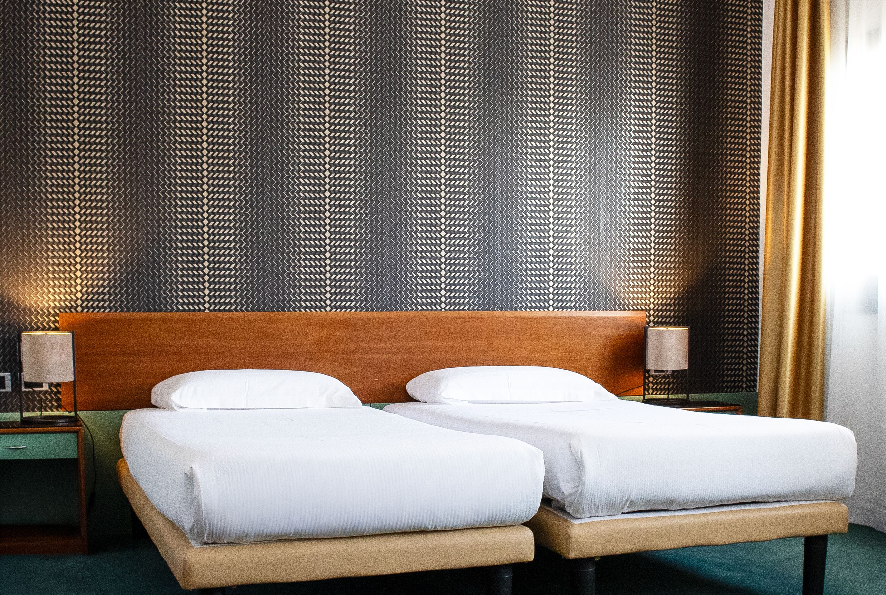 A hotel room with a wooden headboard, two single beds with white linens, a nightstand with a lamp, and a patterned wall.