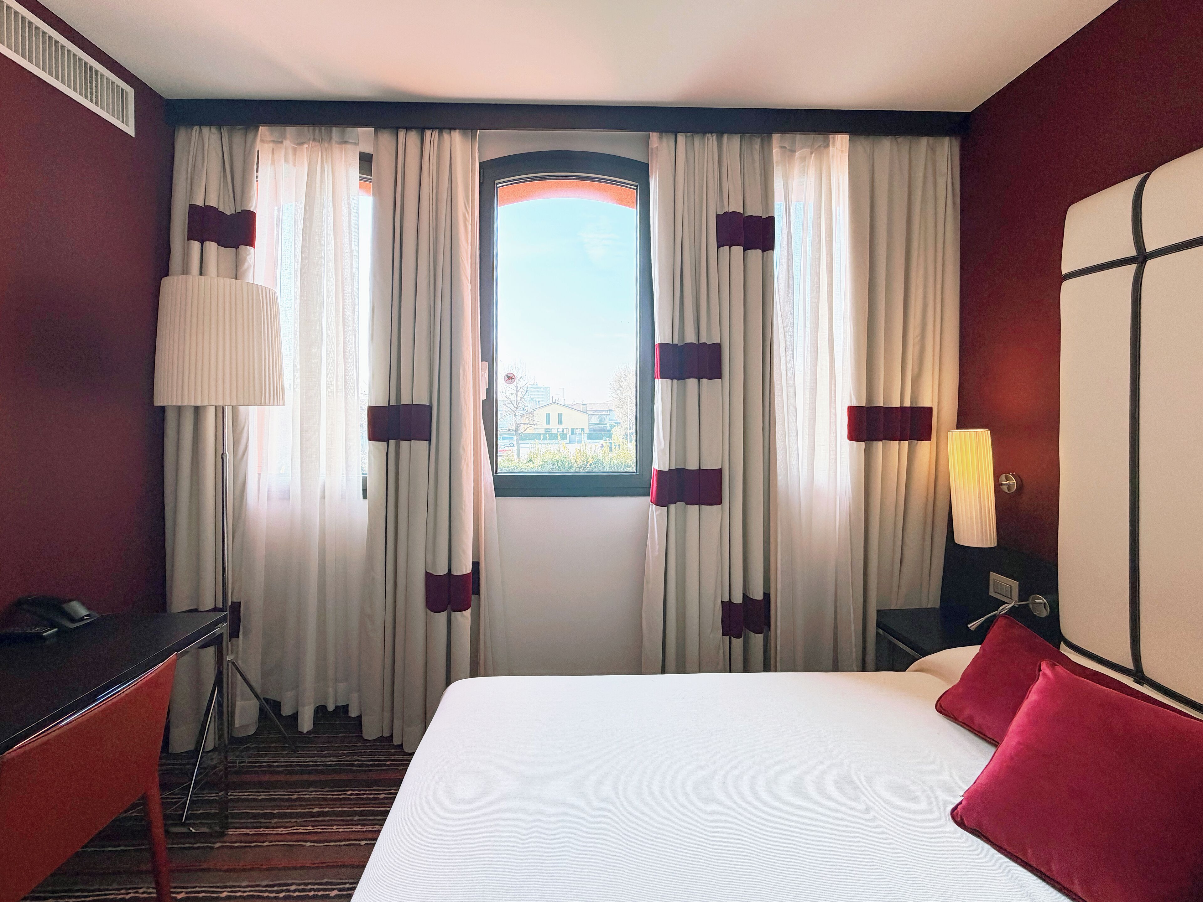 Una habitación de hotel con cama, escritorio, silla, lámpara y cortinas.