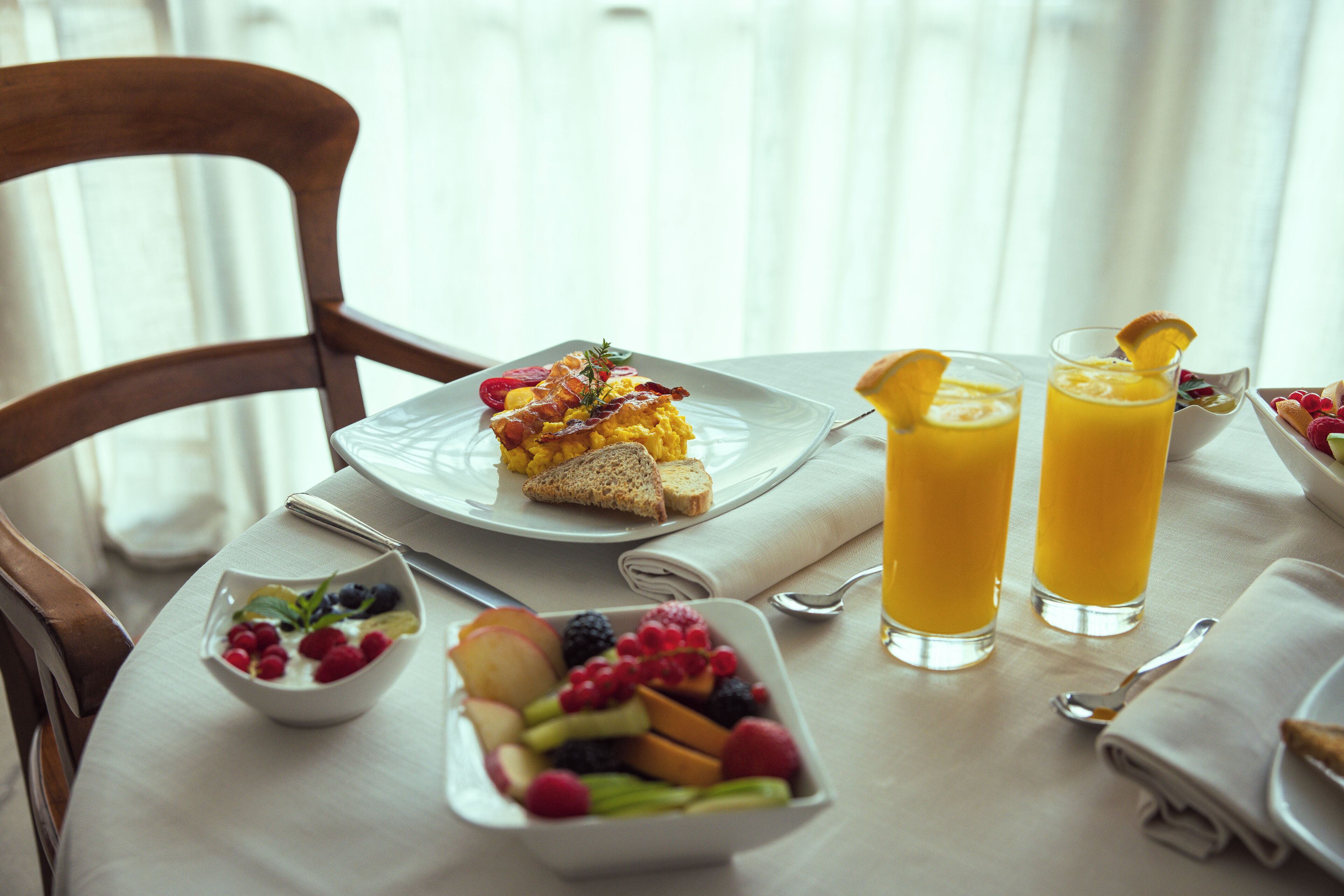 Desayuno con huevos revueltos, tostadas y fruta, dos vasos de jugo de naranja y un tazón de bayas y yogur.