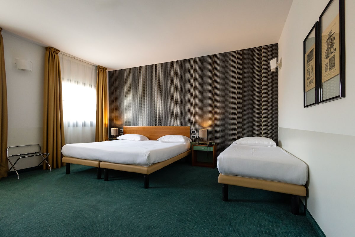 A hotel room with two beds, a green carpet, a wooden headboard, a nightstand with a lamp, and a wall with a textured pattern.