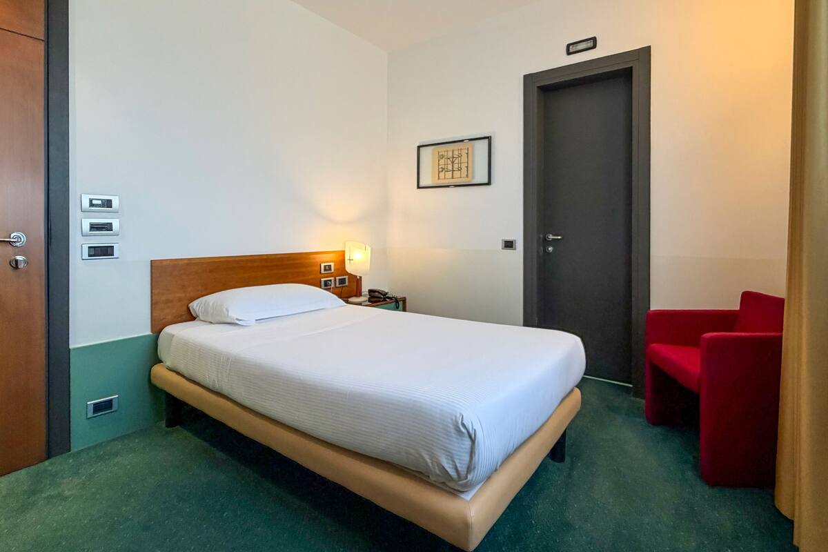 A hotel room with a bed, bedside table, lamp, and a red chair.