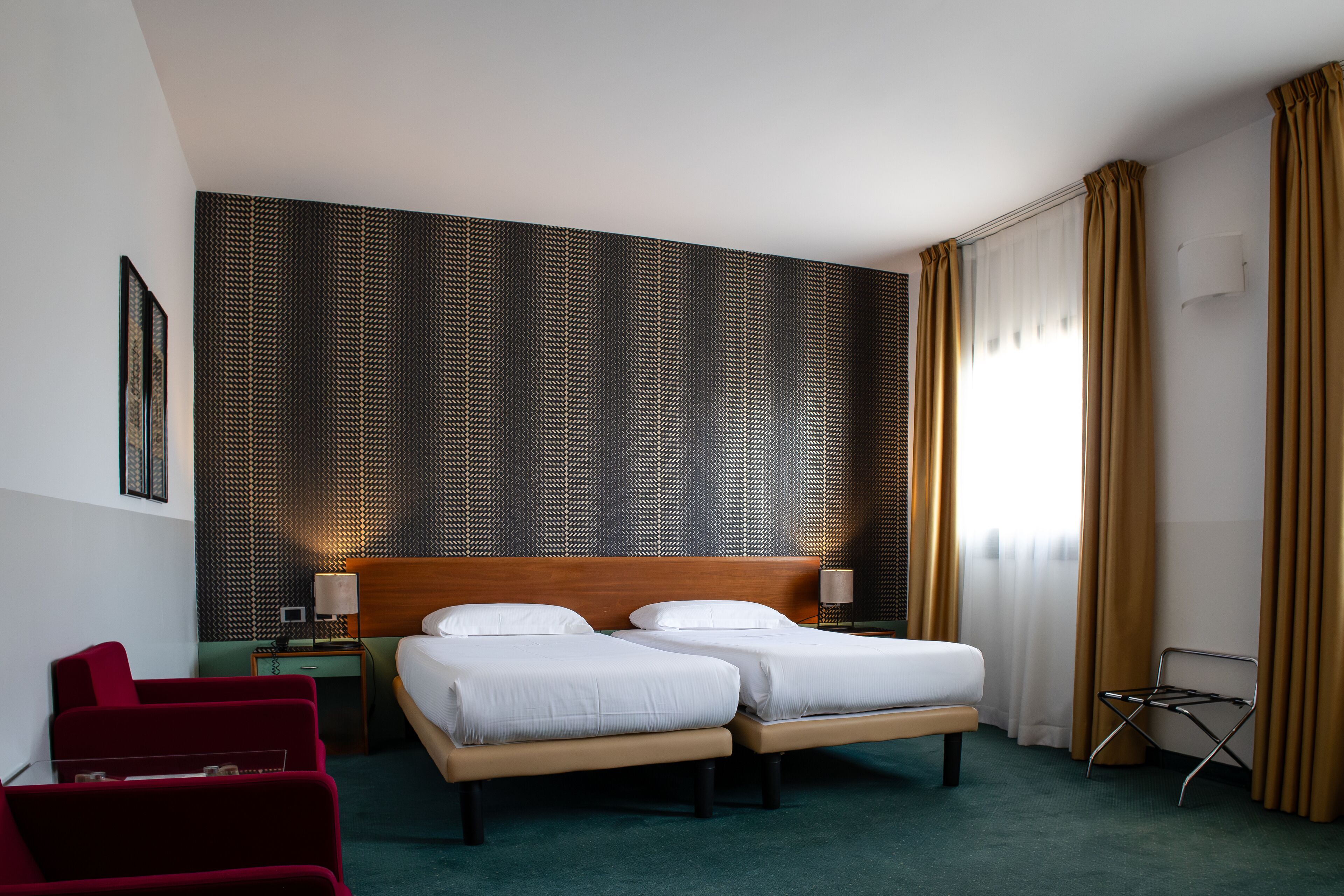 Habitación de hotel con dos camas, un sillón rojo, una mesita con lámpara y una pared con textura.