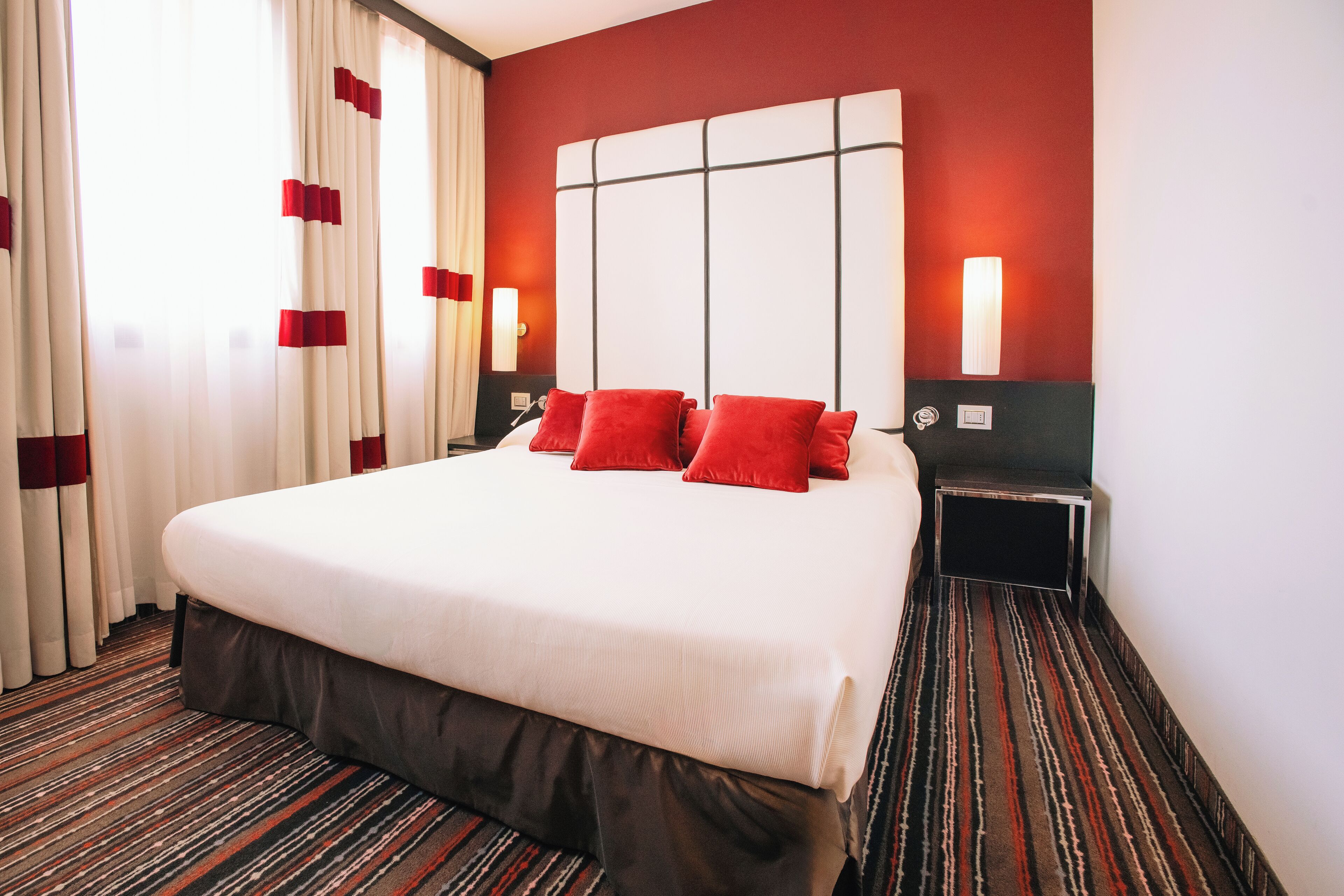 Habitación de hotel con una cama grande, almohadas rojas, cabecera, dos mesitas de noche y alfombra a rayas.