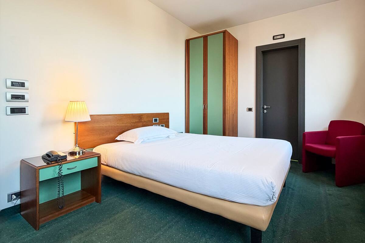 A hotel room with a bed, bedside table, lamp, and a red chair.