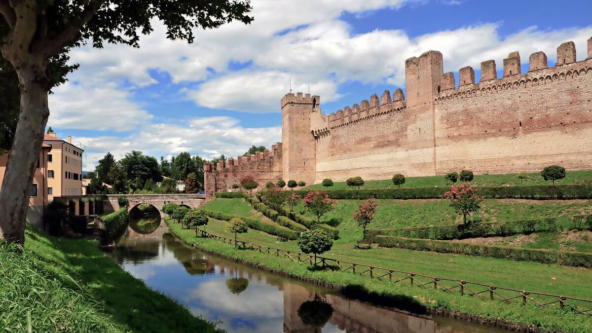 A historic castle with a moat and a bridge, surrounded by well-maintained gardens and a clear sky.