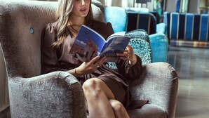 A woman seated in a large armchair reading a book in a well-lit room with a modern interior and a view of the outside through large windows. - Hotel Filanda (Cittadella)
