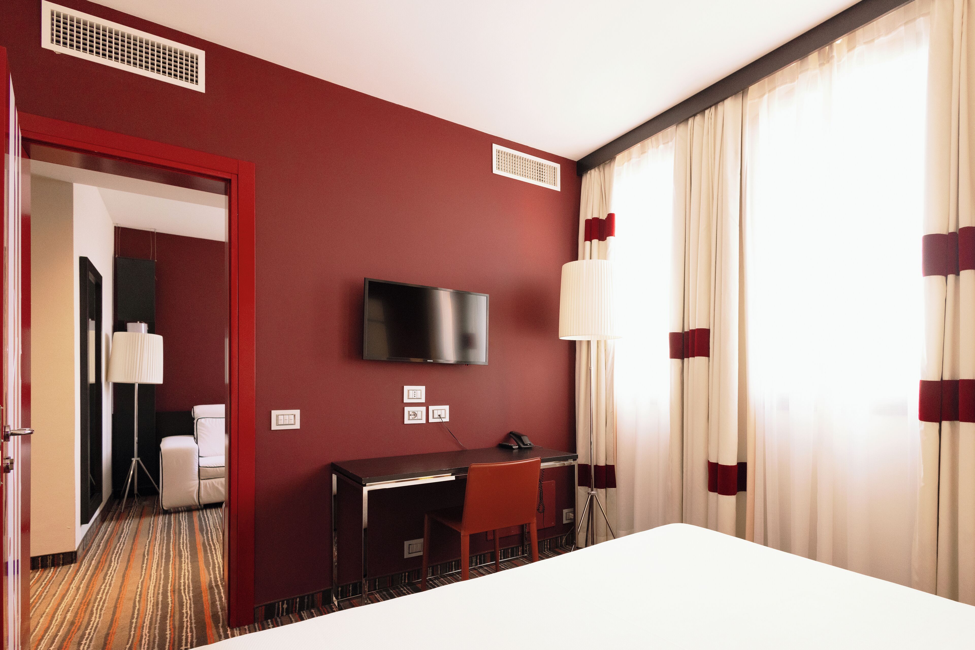 Habitación de hotel con pared roja, televisor de pantalla plana sobre un escritorio, una silla y una cama.