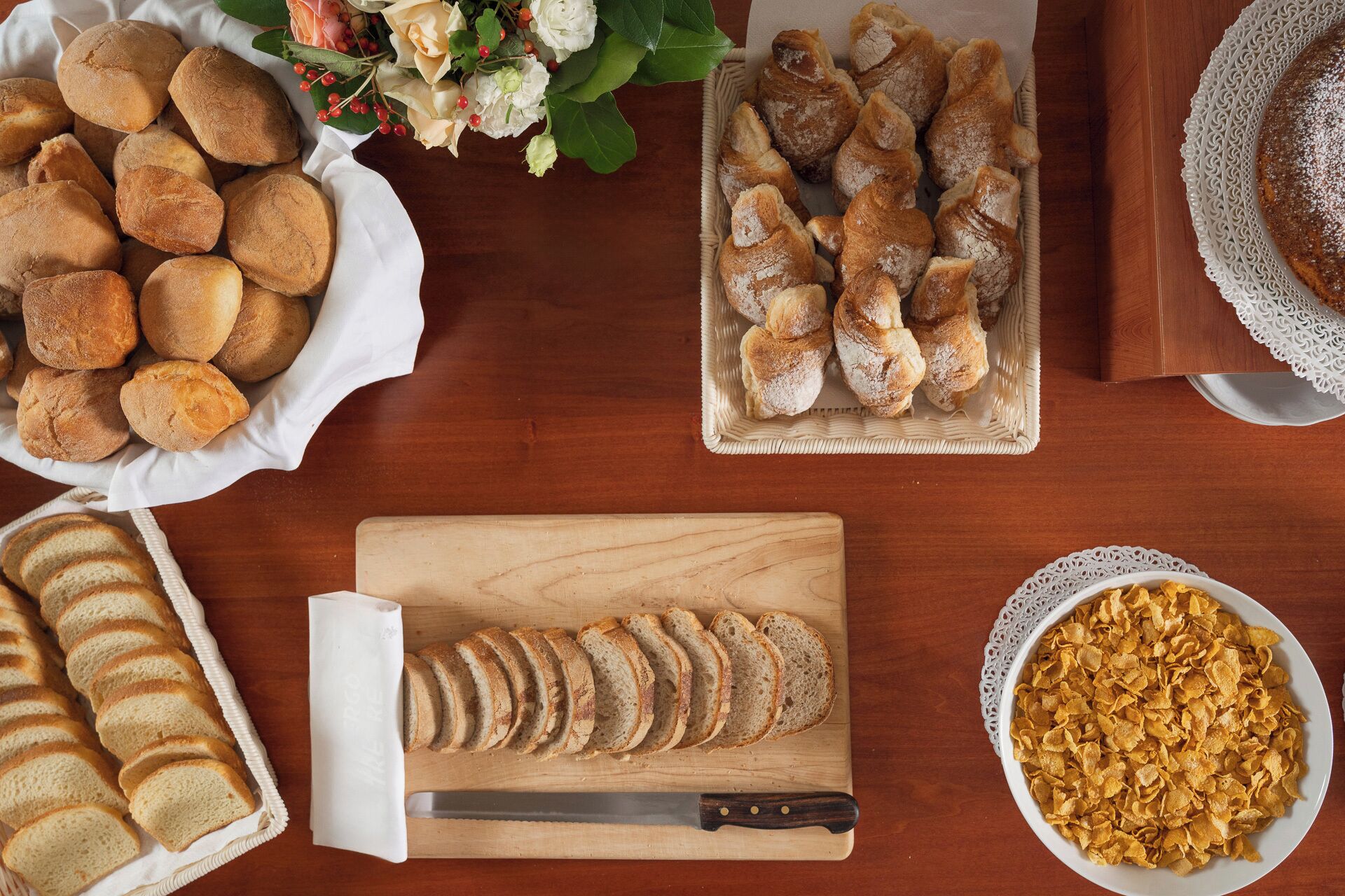 A wooden table with various food items including bread rolls, sliced bread, and cereal.