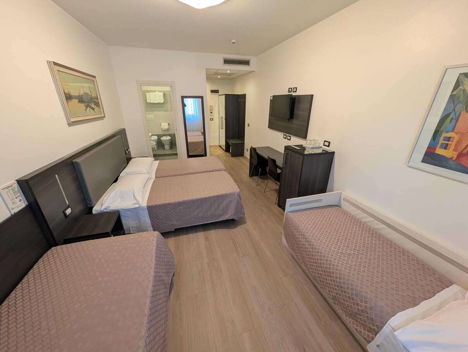 A hotel room with three single beds, a desk, a chair, a TV, and a bathroom visible through an open door.