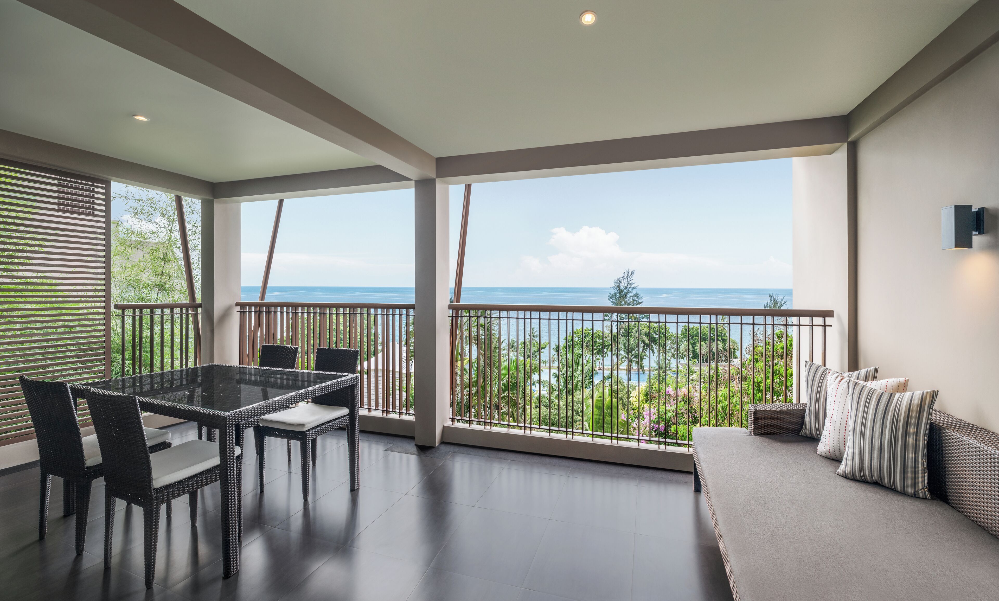 A spacious balcony with a view of the sea, a dining area with wicker furniture, and a seating area with cushions.