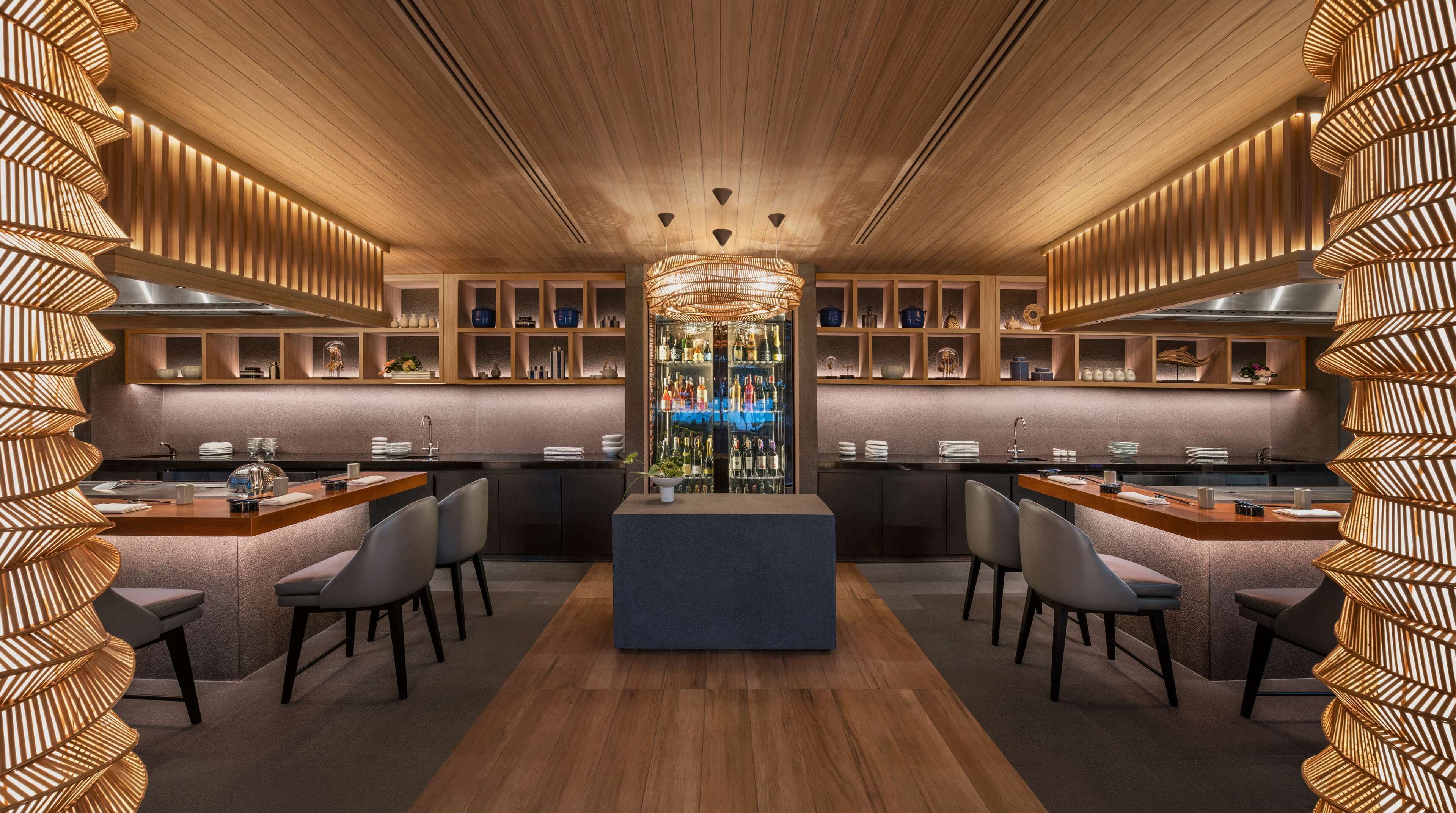 A modern bar area with a central island, wooden paneling, and a variety of bottles and glasses on shelves.