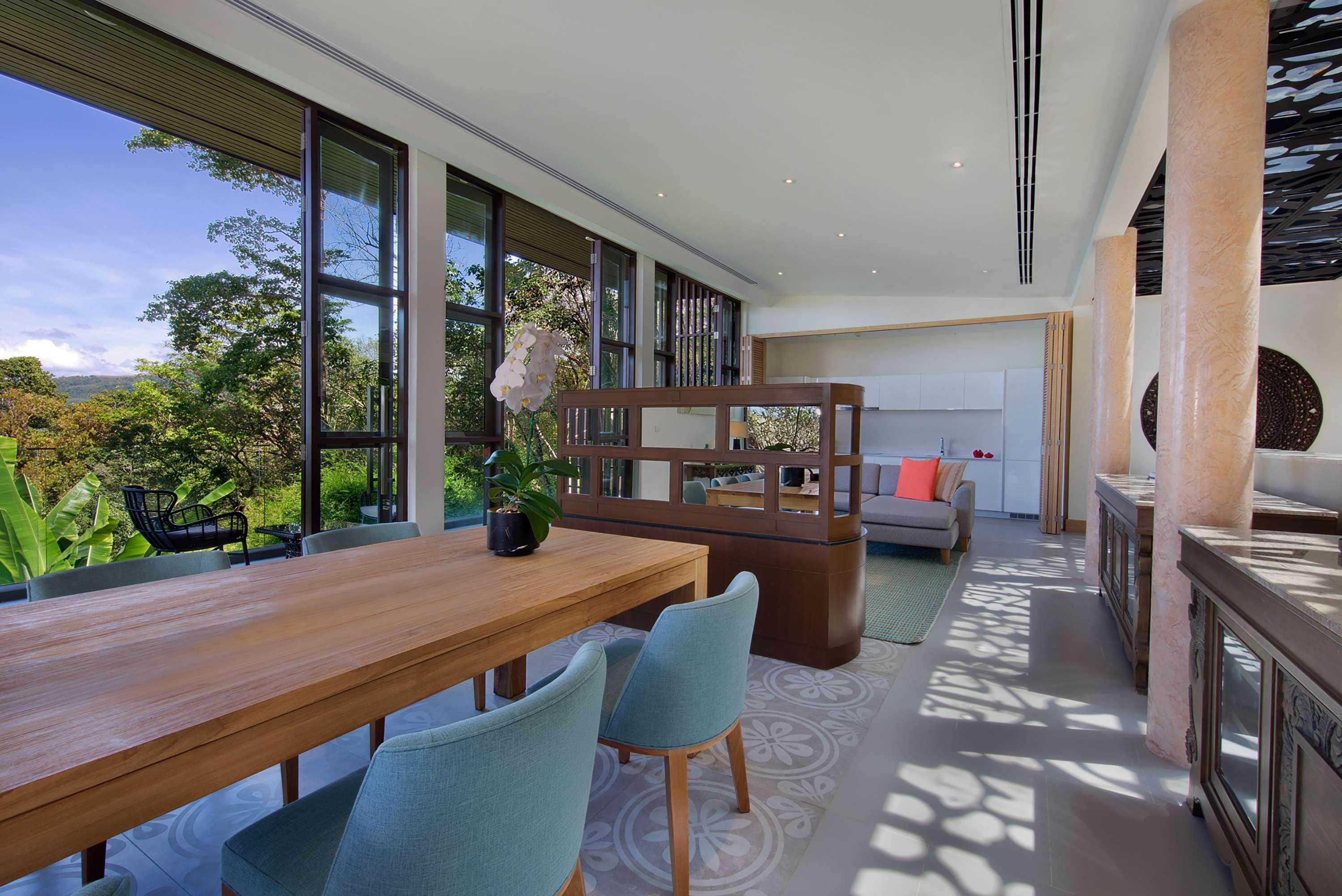 A modern interior with a wooden dining table, two chairs, a sofa, and a large window overlooking greenery.
