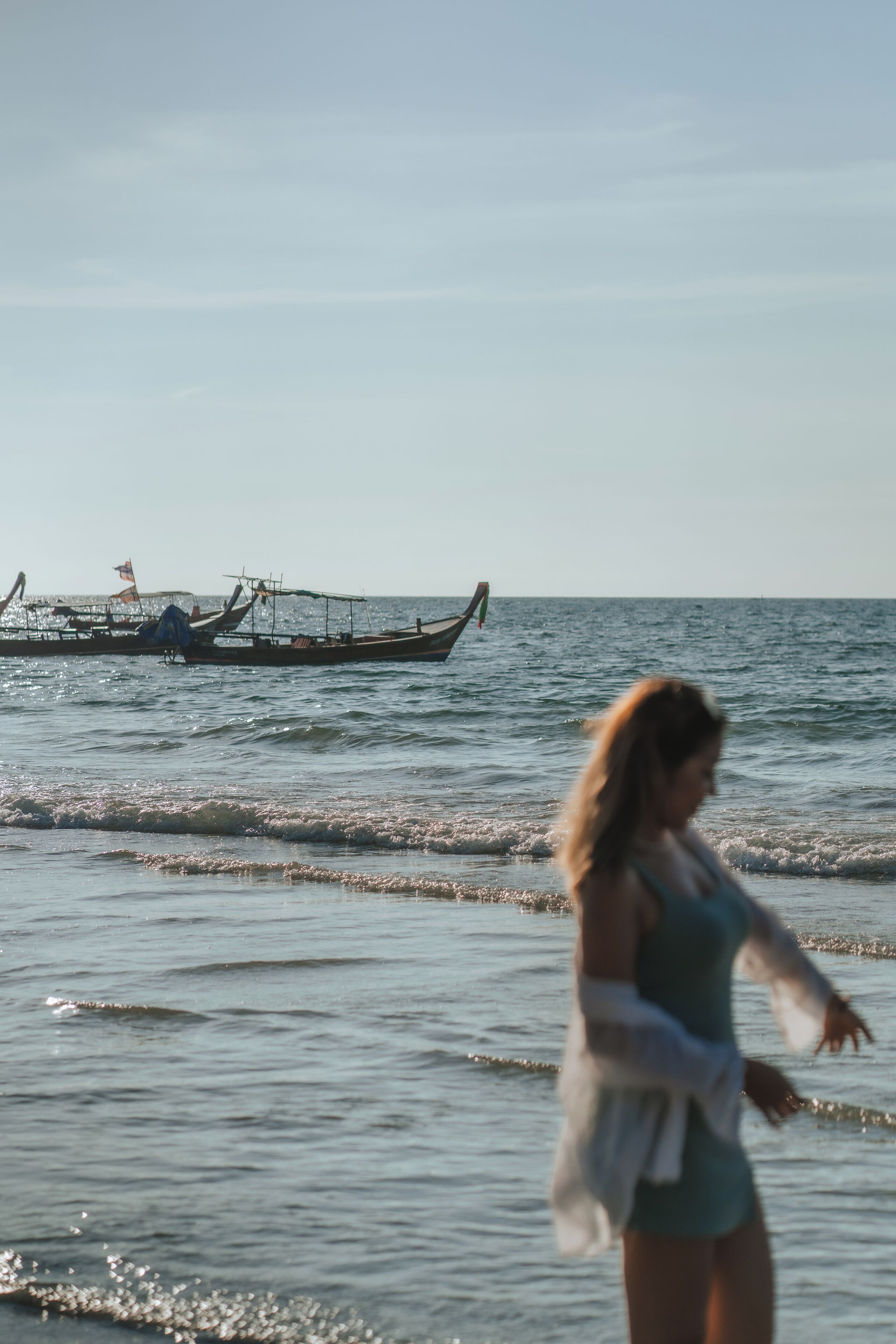 Una persona caminando en la playa con barcos al fondo.