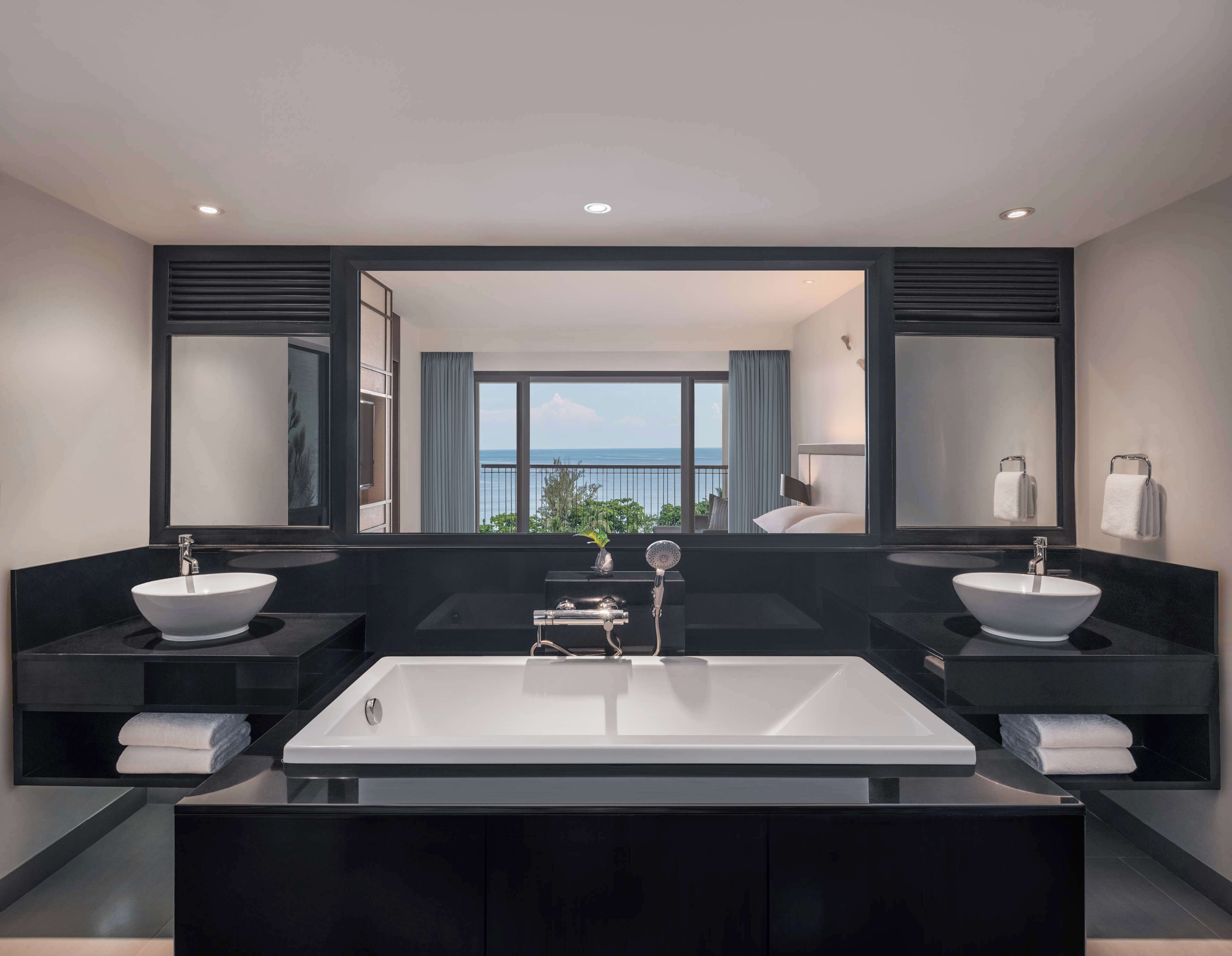 Un baño moderno con dos lavamanos, una bañera grande y vista al mar a través de un ventanal.