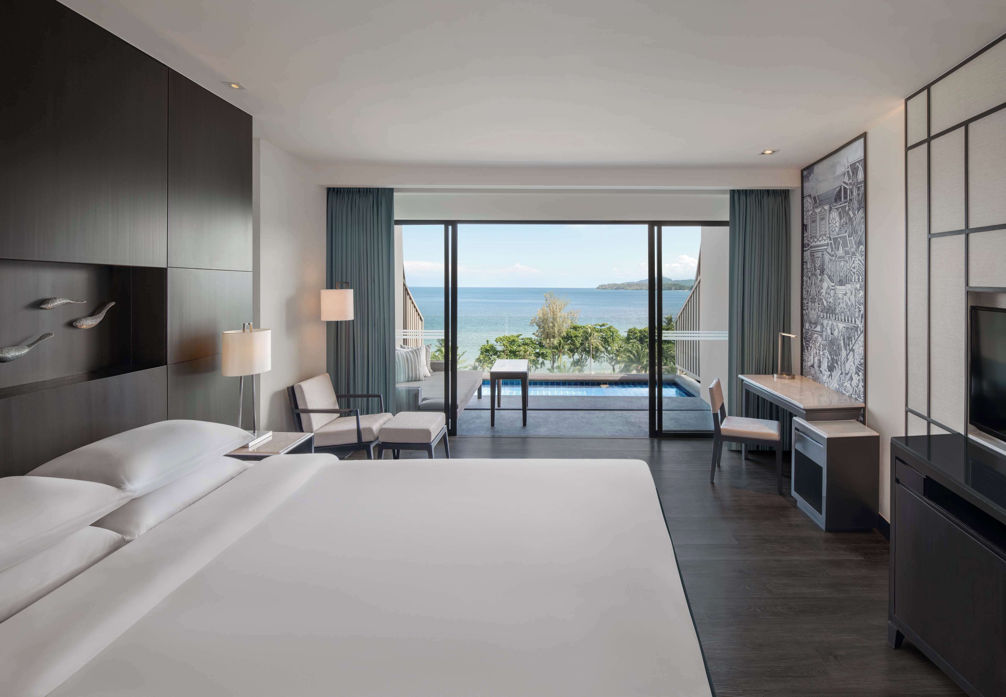 A modern hotel room with a large bed, a desk, a chair, and a view of the sea.