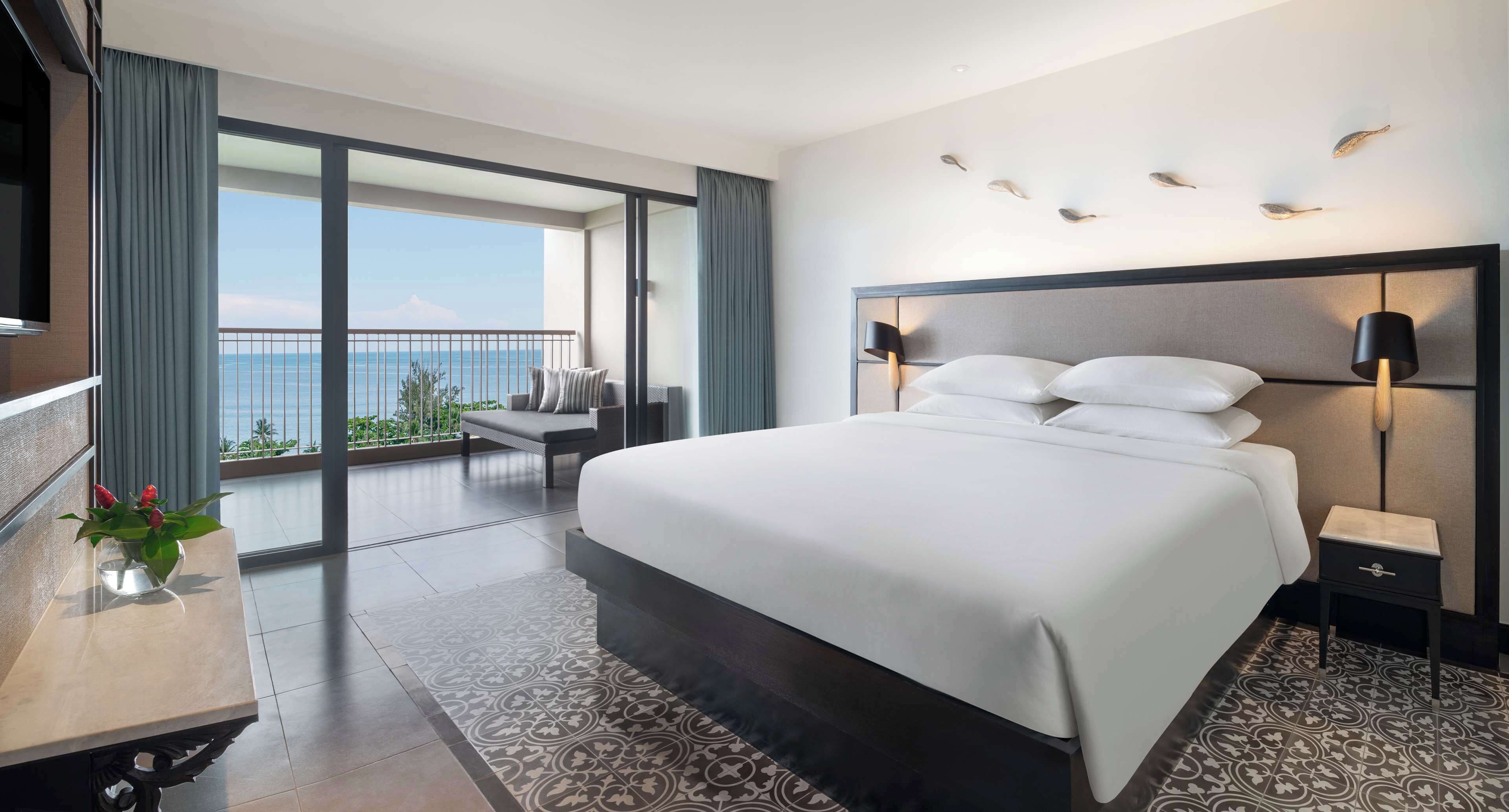 Une chambre d’hôtel moderne dotée d’un grand lit, d’une table de chevet, d’un bureau agrémenté d’un vase de fleurs, d’une vue sur la mer et d’un balcon avec une chaise et une table.